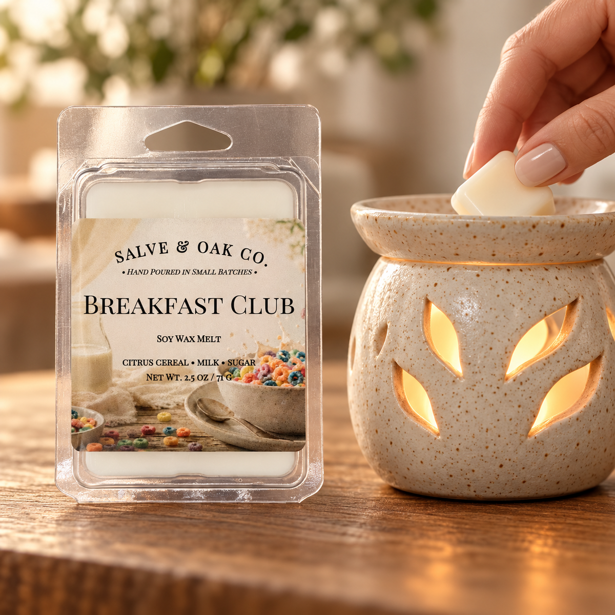 Hand placing an ivory Breakfast Club wax cube into a glowing ceramic warmer beside its package.