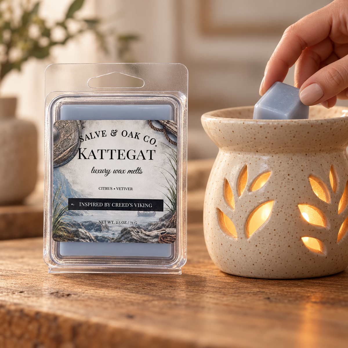 Hand placing a blue-gray Kattegat wax cube into a glowing ceramic warmer beside its package.